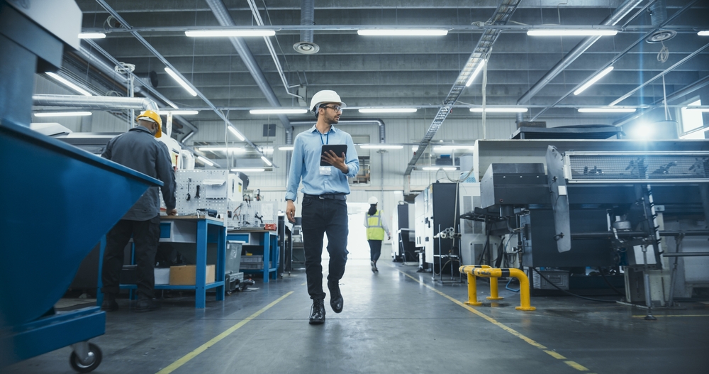 A man in a hard hat walking through a busy factory floor, representing manufacturing operations and cashflow continuity.