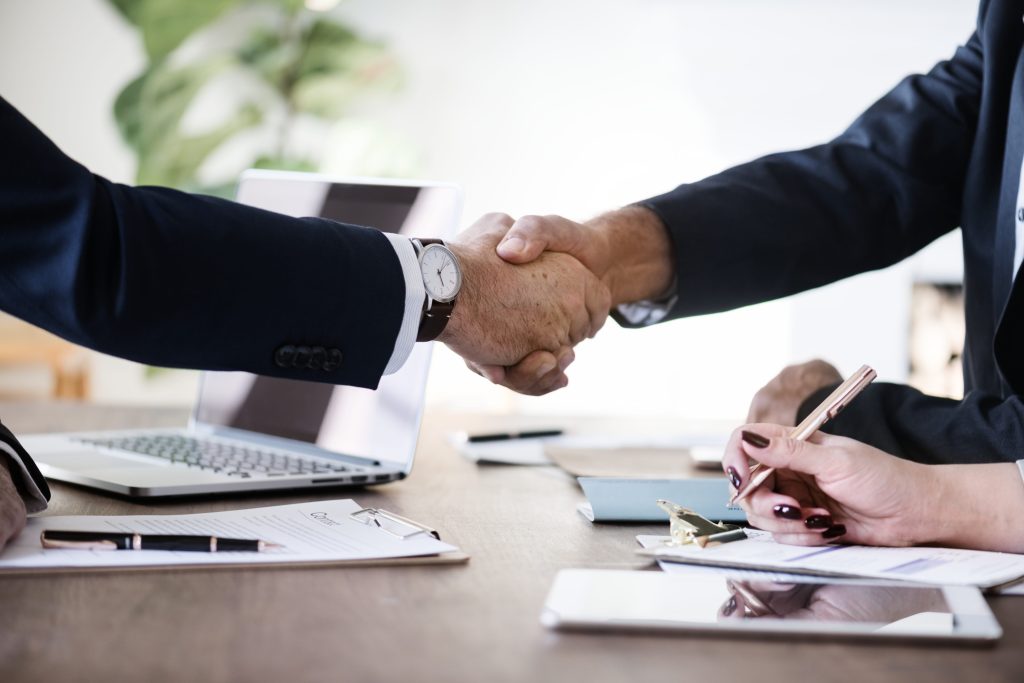Two people in business attire shaking hands across a desk with documents, a laptop, and a tablet.