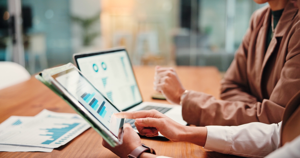 Hands pointing to charts on a tablet while another person works on a laptop displaying financial graphs in a meeting setting.